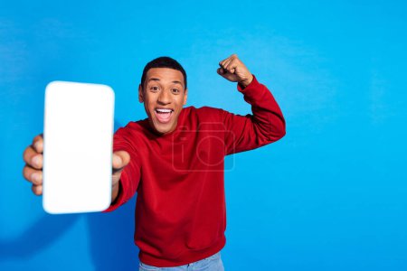 Photo of handsome crazy person showing empty space smart phone screen raise fist isolated on blue color background.