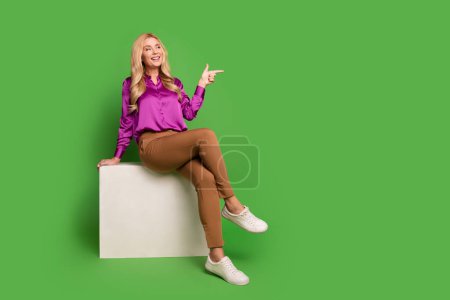 Full length photo of excited thoughtful woman wear violet shirt sitting white platform showing empty space isolated green color background.