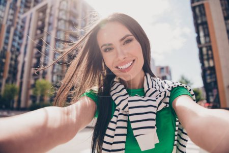 Selfie cadre of video blogger young brunette hair woman wear green t shirt hold camera and toothy smiling on residential complex background.