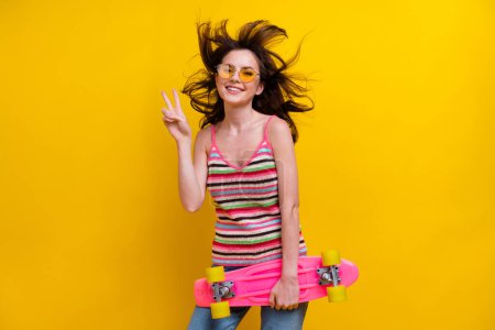 Foto de alegre agradable mujer vestida de punto superior en gafas de sol mantenga monopatín mostrar v-signo aislado en el fondo de color amarillo vibrante.