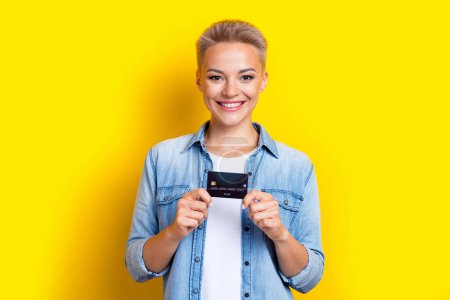 Foto-Porträt der attraktiven jungen Frau halten Kreditkarte Bankkunde tragen trendige Jeans Kleidung isoliert auf gelbem Hintergrund.