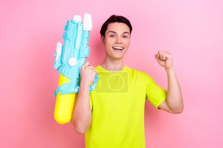 Photo portrait of pretty teenager guy water gun raise fist winning wear trendy yellow outfit isolated on pink color background.