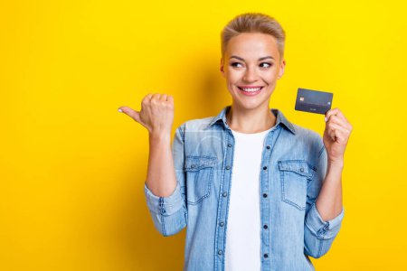 Photo of attractive young woman hold credit card point look empty space dressed stylish denim clothes isolated on yellow color background.