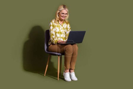 Pleine longueur du corps photo de la femme âgée assise tabouret lecture des rapports quotidiens à l'aide d'un ordinateur portable de bonne humeur isolé sur fond de couleur kaki.