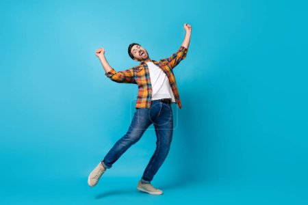 Full body photo of attractive young man dancing raise fists celebrate wear trendy plaid clothes isolated on blue color background.