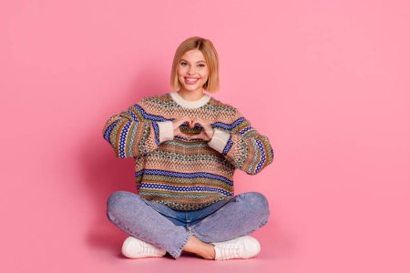 Full length photo of grateful lovely girl dressed print sweater sitting showing heart symbol on chest isolated on pink color background.