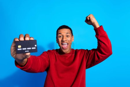 Photo of overjoyed cheerful man wear stylish clothes raise hand fist hold debit plastic card shout yeah isolated on blue color background.