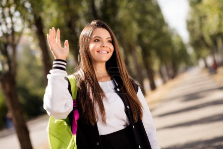 Retrato de agradable chica amigable con cabello moreno llevar elegante mochila uniforme en el hombro decir hola a compañero de clase cerca de la escuela al aire libre.