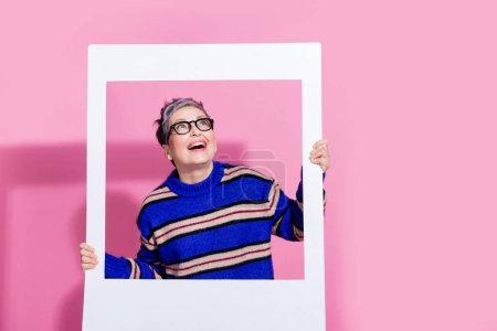 Photo portrait of lovely senior lady look empty space photo frame wear trendy blue striped garment isolated on pink color background.