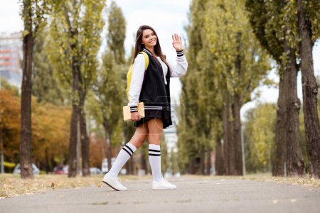 Full length photo of polite girl wear school uniform long socks rucksack go away with book waving you palm on green city street outdoors.