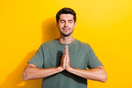 Portrait of peaceful calm positive guy with stubble wear stylish t-shirt put hands together to pray isolated on yellow color background.