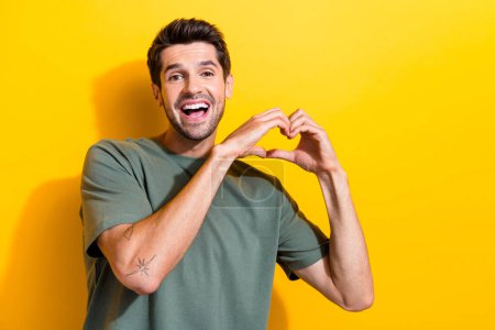 Portrait of cheerful thankful guy with stubble wear stylish t-shirt showing heart symbol smiling isolated on yellow color background.