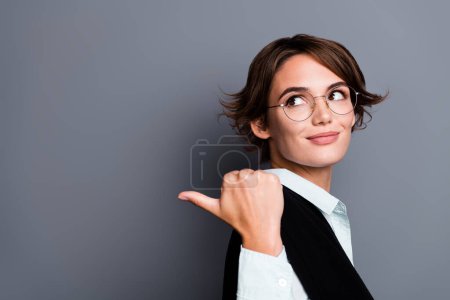 Foto von verträumten positiven Dame ceo gekleidet schwarze Weste Brille suchen zeigt Daumen zurück leeren Raum isoliert graue Farbe Hintergrund.