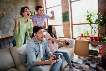 Foto de grupo mejores amigos sentarse sofá jugar competir video juego levantar puños apoyo luz del día plana en el interior.