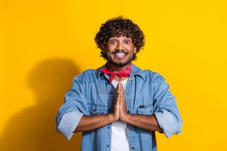 Photo portrait of attractive young man pray hands gesture wear trendy denim clothes red scarf isolated on yellow color background.