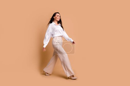 Full length photo of sweet adorable lady dressed white shirt walking heels looking empty space isolated beige color background.