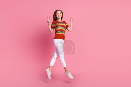 Full body portrait of lovely cheerful person jump demonstrate v-sign empty space isolated on pink color background.