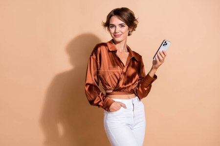 Photo of sweet shiny lady dressed brown shirt texting modern device empty space isolated beige color background.