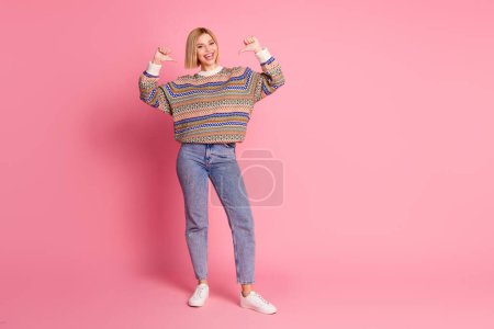 Foto in voller Größe von selbstbewussten schönen Frau tragen Ornament Pullover Jeanshose richtet sich an sich selbst isoliert auf rosa Hintergrund.