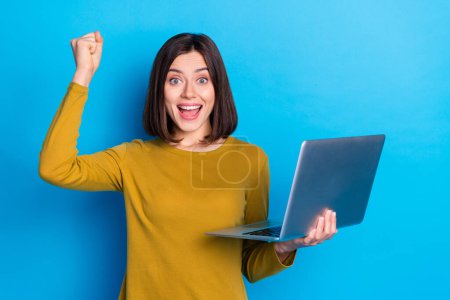 Photo of shocked lucky woman wear shirt rising fist winning game modern device empty space isolated blue color background.