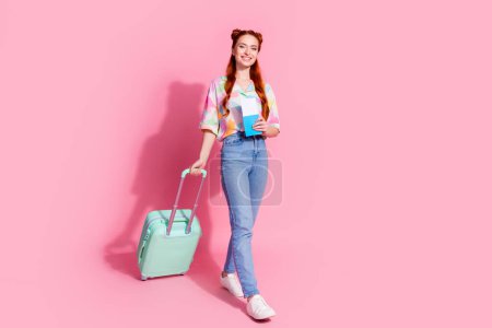 Full size photo of pleasant girl dressed colorful blouse jeans pants walk with valise hold tickets isolated on pink color background.