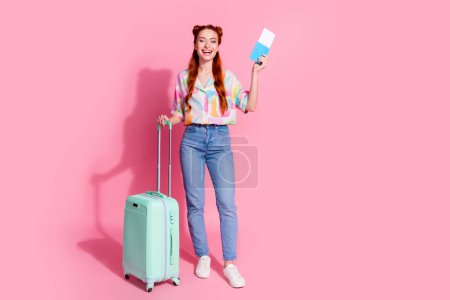 Full size photo of satisfied girl dressed colorful blouse jeans pants stand with valise hold tickets isolated on pink color background.