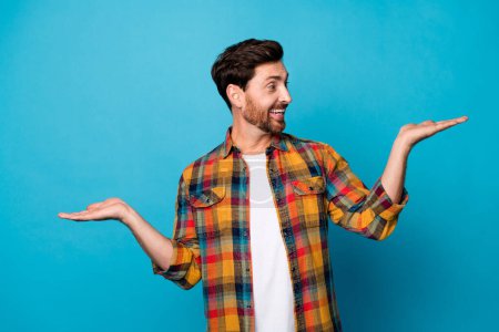 Photo of satisfied man with beard dressed plaid shirt look at better product on palm empty space isolated on blue color background.