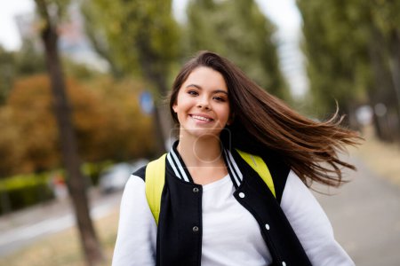 Photo de toothy rayonnant étudiant avec des battements droits sur les cheveux du vent habillé sac à dos veste élégant retour à l'école en automne à l'extérieur.