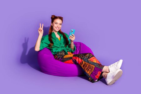 Foto in voller Länge von fröhlichen Mädchen tragen Print Vintage-Hosen sitzen auf Hocker halten Smartphone zeigt V-Zeichen isoliert auf violettem Hintergrund.