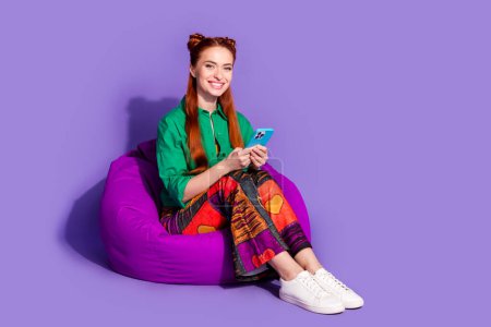 Ganzes Foto von optimistischen Mädchen tragen Print Vintage-Hosen sitzen auf Hocker halten Smartphone chattet isoliert auf violettem Hintergrund.