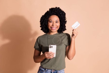 Photo of good mood nice woman wear khaki t-shirt holding smartphone demonstrate debit card in arm isolated on beige color background.