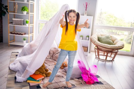 Photo portrait of adorable small girl show thumbs up good playtime tent dressed stylish yellow garment preschoolers kindergarten interior.