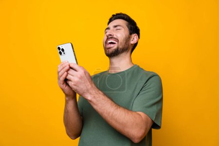 Photo portrait of handsome young guy hold telephone laugh funny joke dressed stylish khaki outfit isolated on yellow color background.