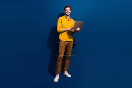 Foto in voller Länge von netten jungen Mann halten Netbook-Arbeit aus der Ferne gekleidet stilvolle gelbe Kleidung isoliert auf dunkelblauem Hintergrund.