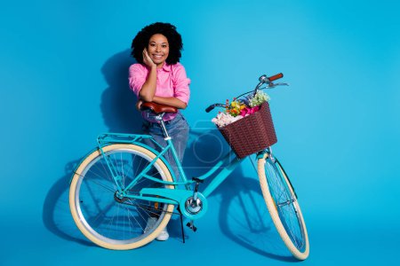 Full length photo of optimistic girl dressed silk shirt jeans lean on bike keep arm on cheekbone isolated on blue color background.