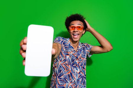 Photo of impressed excited guy wear print shirt dark eyeglasses showing you modern device empty space isolated green color background.