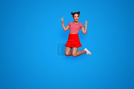 Full-length full-size view of jumping laughing and pretty woman dressed in colourful bright clothes shows a v-sign isolated on red background. Joy fun concept.