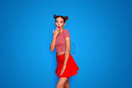 Portrait of cute and surprise girls dressed in striped tshirt and red skirt covers her open mouth with palm isolated over background.