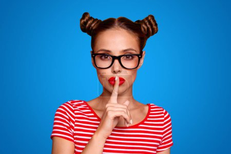  Portrait of attractive mysterious girl in glasses gesturing silence sign with forefinger red pout lips looking at camera isolated on red background.