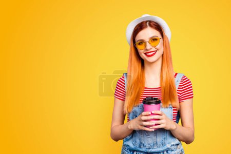 Cute young girl looks into the camera and holds pink paper glass with a drink in her hands isolated on bright blue background with copy space for text.