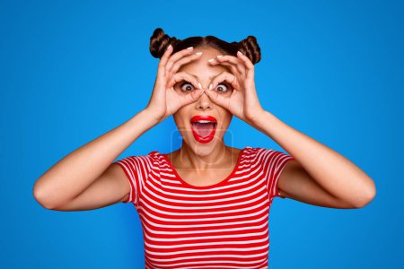 Pretty girl with surprised face holding fingers near eyes like glasses: mask like super hero or owl isolated on red background.