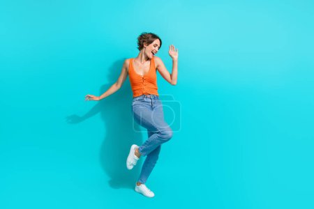 Full length photo of good mood carefree cute woman wear orange singlet jeans dancing having fun isolated on turquoise color background.