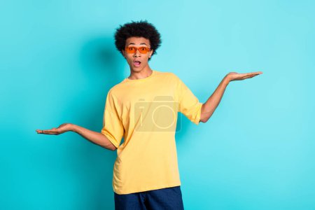 Photo of confused guy with afro hair dressed yellow t-shirt in sunglass palms comparing products isolated on turquoise color background.