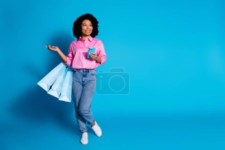 Full length portrait of pretty young woman hold phone bags empty space wear pink shirt isolated on blue color background.