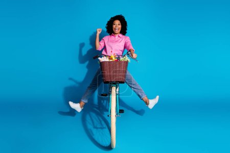 Foto de longitud completa de la chica agradable extática vestida camisa de seda vaqueros montar bicicleta apretando puño divertirse aislado sobre fondo de color azul.