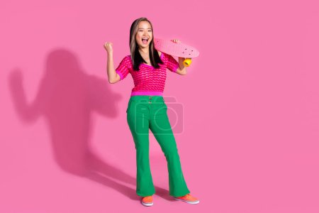 Foto de longitud completa de la dama de la suerte fresco vestido camisa de punto monopatín levantamiento puño vacío espacio aislado color rosa fondo.