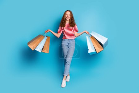 Foto de tamaño completo de hermosa chica usar elegantes pantalones de mezclilla camiseta sosteniendo bolsas de compras en las manos aisladas sobre fondo de color azul.