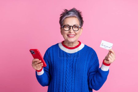Photo de charmante belle femme âgée gaie porter des vêtements bleus tendance tenir carte bancaire payer acheter nfc isolé sur fond de couleur rose.