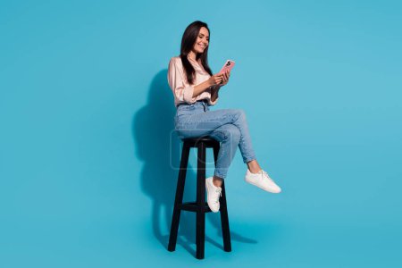 Pleine longueur photo de femme positive gaie porter chemisier soyeux assis tabouret communiquant dispositif moderne isolé fond de couleur bleue.