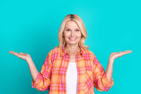 Portrait of positive woman blond hair dressed plaid shirt arms comparing products empty space isolated on turquoise color background.